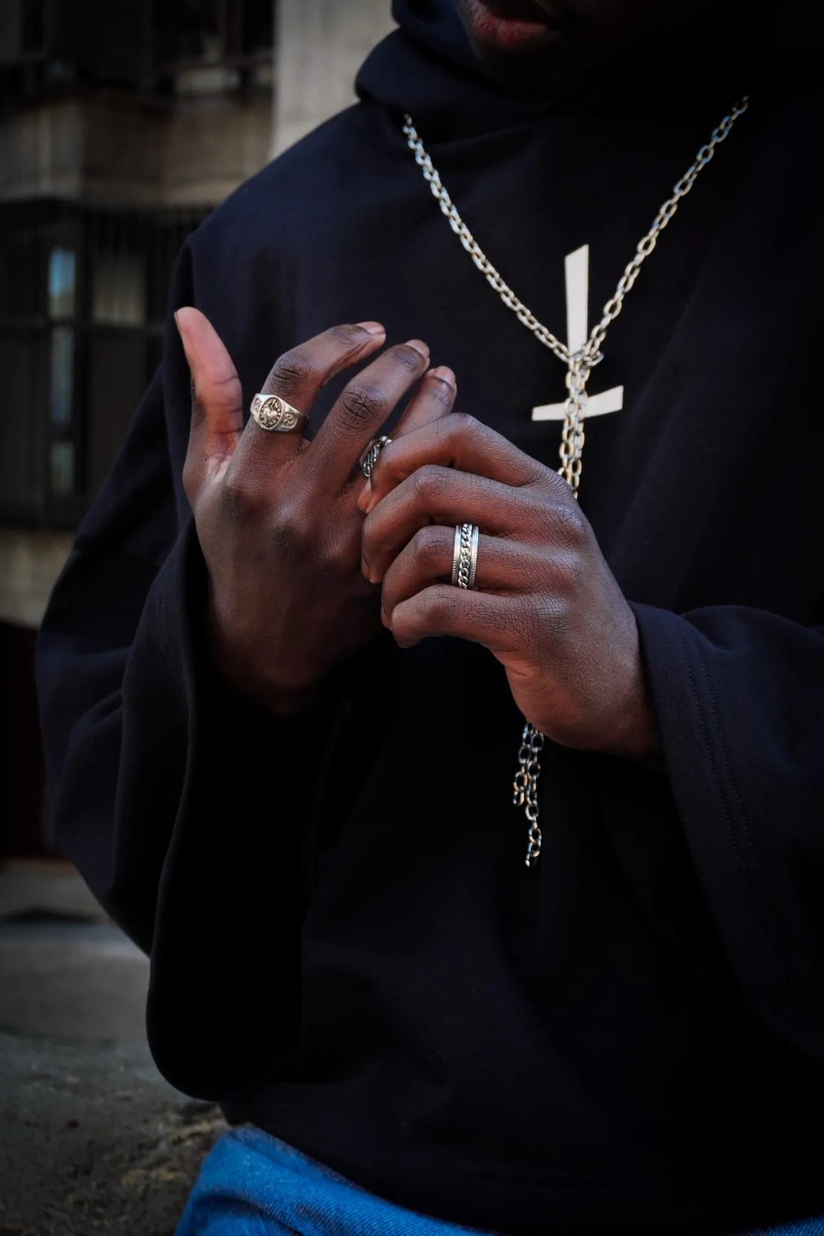 Primer plano de manos con anillos plateados y collar cruz Skull Hearts Drop 02 sobre sudadera negra.